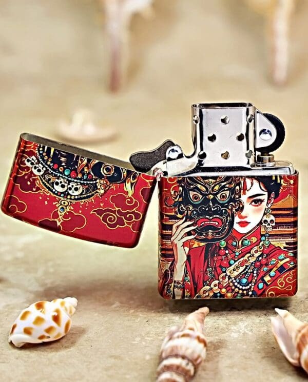 Decorative metal lighter with red Asian-inspired art showing a woman and a demon mask, lid open, on a sandy surface with shells nearby (informational product image).
