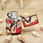 Metal lighter with a red and gold decorative Asian-inspired design, open lid, resting on a sandy surface with seashells nearby