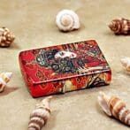 Decorative red tin box with ornate Asian-inspired artwork, placed on sand among seashells.