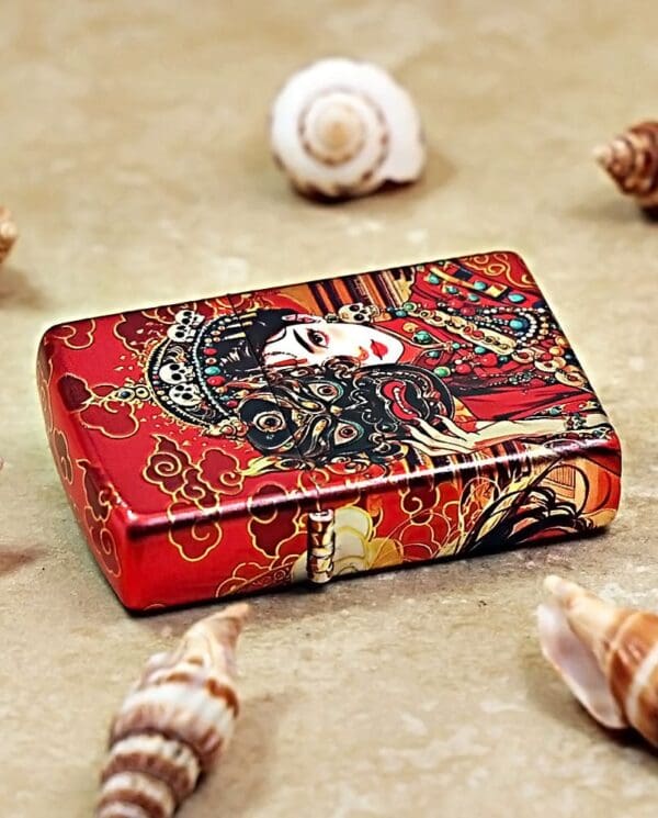 Decorative red tin box with ornate Asian-inspired artwork, placed on sand among seashells.