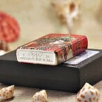 Decorative red Zippo lighter with ornate pattern resting on a black box; metal bottom shows 'Zippo' and 'Made in USA' (Bradford, PA).