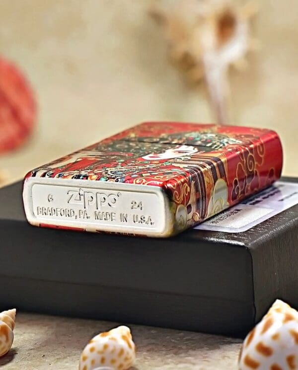 Decorative red Zippo lighter with ornate pattern resting on a black box; metal bottom shows 'Zippo' and 'Made in USA' (Bradford, PA).
