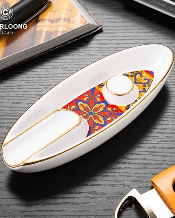 White ceramic boat-shaped ashtray with a colorful floral inlay and gold trim, brand logo visible in the corner.