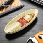 Boat-shaped ceramic ashtray with gold trim and a colorful floral inlay, placed on a dark wood surface with Cigarloong branding in the corner.