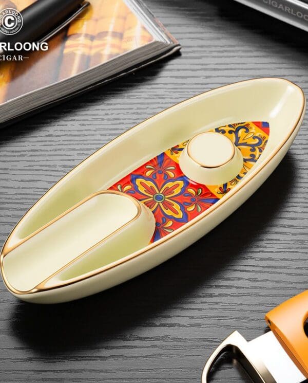 Boat-shaped ceramic ashtray with gold trim and a colorful floral inlay, placed on a dark wood surface with Cigarloong branding in the corner.