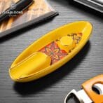 Yellow cigar cutter with a floral inlay on a dark wooden table, beside a cigars box.