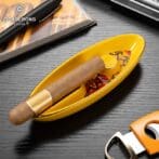 Cigar resting in a yellow oval ashtray on a dark wood table, with a book and cigar cutter nearby.