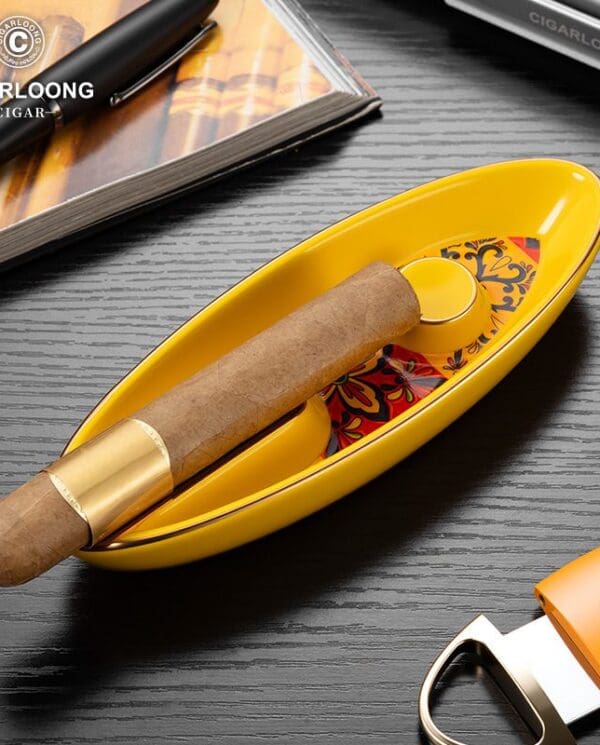 Cigar resting in a yellow oval ashtray on a dark wood table, with a book and cigar cutter nearby.