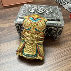 Gold-toned ornate ceremonial mask/helmet figurine with red and teal accents and intricate raised patterns, resting on a wooden table beside a silver decorative box.
