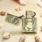 Gold engraved lighter on a sandy beach with seashells, lid open, featuring a bikini-clad woman and floral details in black and gold.