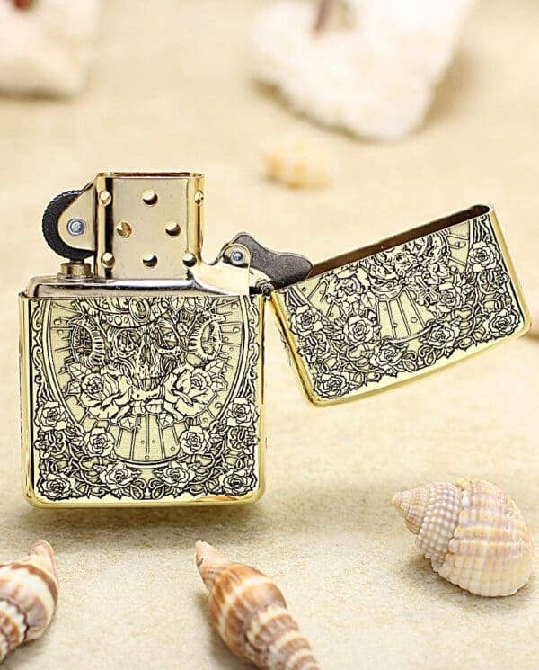 Gold engraved Zippo-style lighter opened, resting on a sandy beach with seashells nearby; vintage floral pattern on the case.