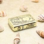 Gold Zippo lighter with ornate floral and figure engraving, set on sand among seashells.