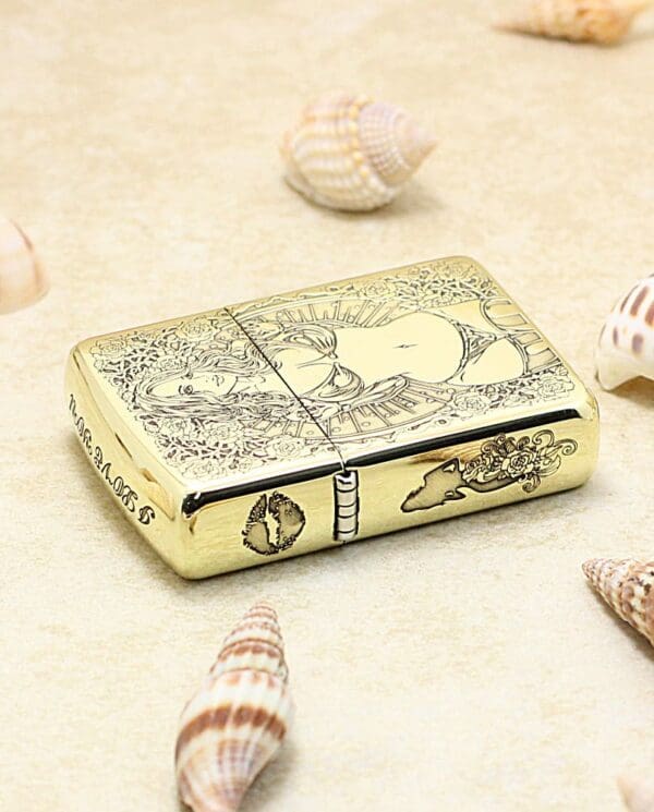 Gold Zippo lighter with ornate floral and figure engraving, set on sand among seashells.